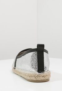 Espadrille argenté avec une texture cuir craquelée, des accents noirs, une forme arrondie et une semelle en jute tressé. La vue arrière montre une bande de traction.