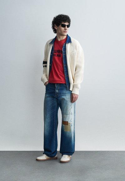 Homme portant des lunettes de soleil, t-shirt rouge à motif superposé d'une veste en jean et d'un cardigan en maille crème, jean large à patchs bleus, baskets blanches.