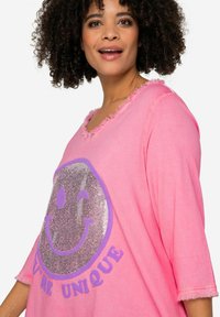 Rosa Oversize-Hemd mit einem ausgefransten V-Ausschnitt, das ein großes glitzerndes Smiley-Gesicht und den Text "DU BIST EINZIGARTIG" in lila darunter zeigt.