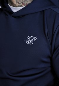 Felpa blu navy realizzata in tessuto liscio, dotata di un logo ricamato bianco sul petto. Il design include un cappuccio e una texture morbida.