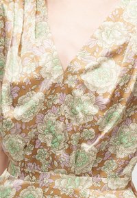 Καφέ φόρεμα με μεγάλο ανοιχτό πράσινο και κρεμ λουλουδάτο μοτίβο, ντεκολτέ σε σχήμα V και εφαρμοστή μέση, εμφανιζόμενο στο πάνω μέρος του κορμού.