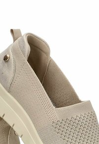 Zapatos beiges sin cordones con una parte superior de tela texturizada, que presenta un patrón de malla. Un pequeño detalle dorado es visible en el lateral. Suela blanda y flexible.