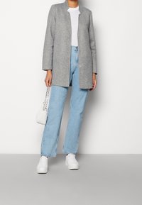 Abrigo gris de manga larga con un diseño minimalista, combinado con jeans de pierna ancha en azul claro y zapatillas blancas. Se incluye un pequeño bolso blanco.