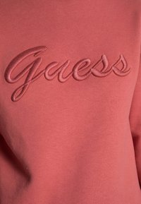 Detailní záběr na růžovou látku s vyvýšeným vyšitým slovem 'Guess' ve stejnobarevné růžové nitě.