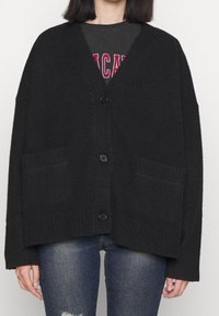 Svart stickad cardigan med långa ärmar, V-ringning, tre knappar och två framsidofickor. Ribbad textur vid fållen och fickkanternas kanter.