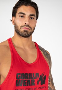 Red tank top made of fabric features a black graphic text "GORILLA WEAR" and a gorilla illustration. The design includes a round neckline and wide armholes.