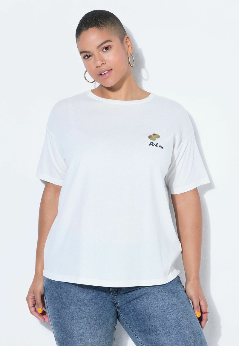 Žena s krátkými vlasy, nosící zlaté kruhové náušnice, bílé tričko s nápisem „Pick me“ a olivovým motivem, a modré džíny, stojící před bílým pozadím.