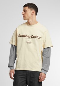 T-shirt beige à manches courtes avec le texte marron "AnotherCotton." Superposé avec un sous-pull gris à manches longues, présenté avec un jean foncé.