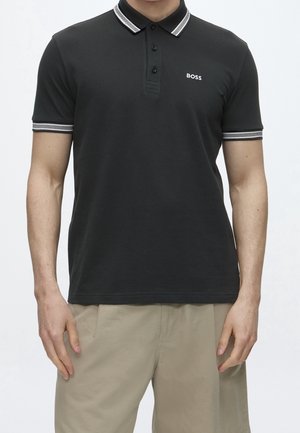 Mann trägt schwarzes Poloshirt mit weiß gestreiftem Kragen und Ärmeln, "BOSS" Logo auf der Brust, kombiniert mit beigen Shorts.