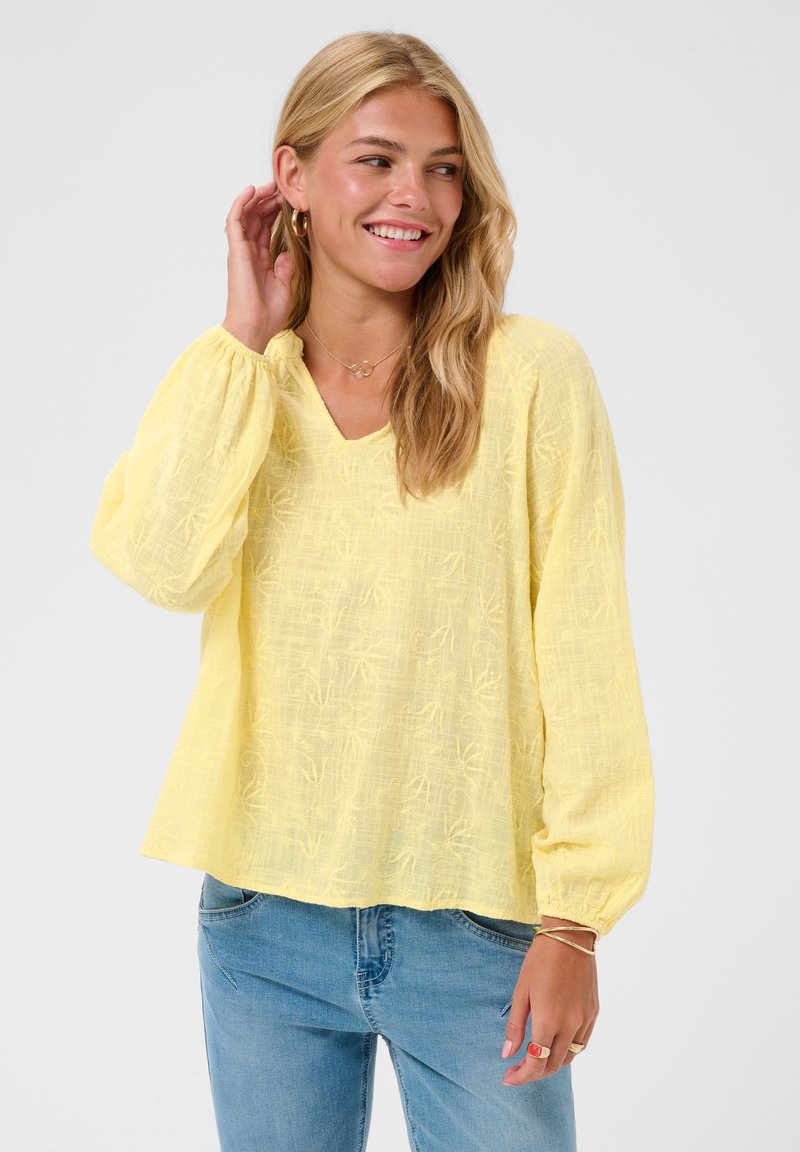 Jeune femme aux cheveux blonds portant un chemisier jaune texturé à manches longues ample et un jean bleu, souriante et se touchant les cheveux