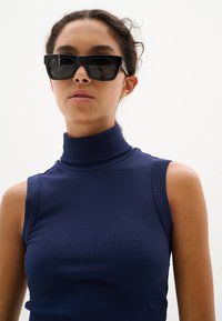 Occhiali da sole neri rettangolari abbinati a un maglione a collo alto blu navy aderente, caratterizzato da una texture a coste e design senza maniche.