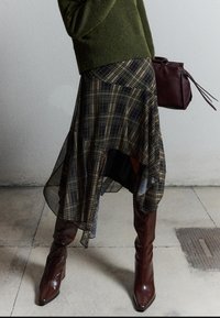 Kvinde iført en grøn sweater, ternet asymmetrisk nederdel, høje brune læderstøvler, holder en mørkerød håndtaske foran en grå væg.