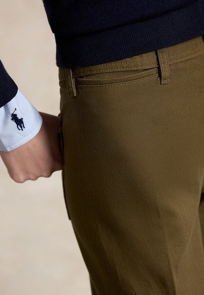 Person trägt olivgrüne Hose und marineblauen Pullover mit weißem Hemdmanschettenaufschlag, der ein marineblaues Polo-Spieler-Logo zeigt.