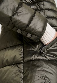Olijfgroene pufferjas met een doorgestikte textuur, voorzien van zwarte knopen en een ritssluitingzak. Heeft een geribbelde witte mouwdetail.