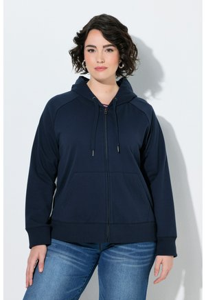 Kvinde med kort krøllet hår iført sølv hoops, en mørkeblå lynlåshoodie og blå jeans, stående mod en hvid baggrund.