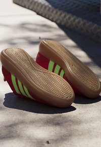 Pair of red sneakers with green stripes showing textured beige soles, placed on sunlit pavement next to a shadowed edge.