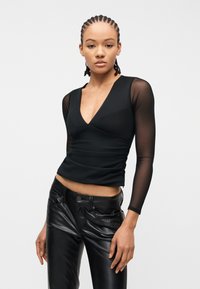 Νεαρή γυναίκα με πλεξούδες, που φοράει μαύρο τοπ με V λαιμόκοψη και διαφανή μακριά μανίκια καθώς και μαύρο δερμάτινο παντελόνι, στέκεται μπροστά σε ένα απλό φόντο.
