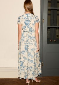 Rochie maxi din material bej deschis cu imprimeu de peisaj bleu, maneci scurte, corset ajustat și fustă fluida. Legată la talie, design minimalist.