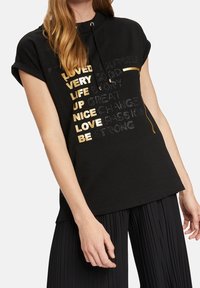 Sudadera negra de manga corta con capucha y cordón. Presenta letras texturizadas en dorado y plateado con frases motivacionales. Tejido suave, ajuste relajado.