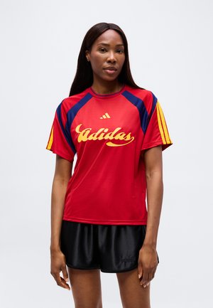 Žena má na sebe červené športové tričko Adidas so žltým logom a tmavomodrými prvkami, v kombinácii s čiernymi šortkami Adidas, stojí pred jednoduchým pozadím.