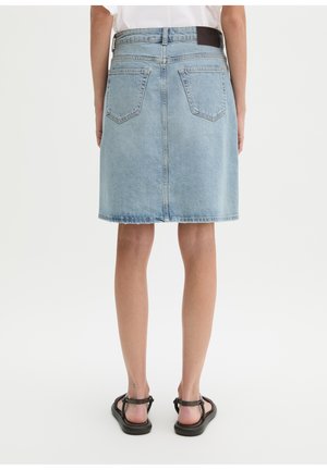 Personne portant une jupe en denim bleu clair jusqu'aux genoux avec des poches à l'arrière et des sandales noires, debout devant un fond blanc uni.