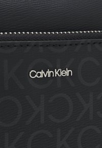 Black synthetic wallet featuring embossed Calvin Klein logo, silver hardware, smooth texture, and a zip closure along the top edge.