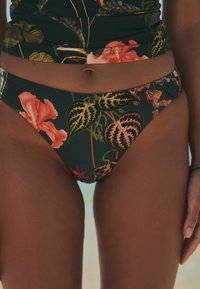 Cvetni bikini spodnji del z temno zeleno podlago in velikimi rožnatimi in rdečimi hibiskusi ter listnatimi vzorci, ki poudarjajo prilegajoč dizajn.
