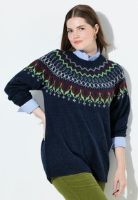 Maglione lavorato a maglia blu navy con un colletto decorato da un motivo colorato in rosso, verde e bianco, indossato sopra una camicia colletto azzurro. Design a vestibilità larga.