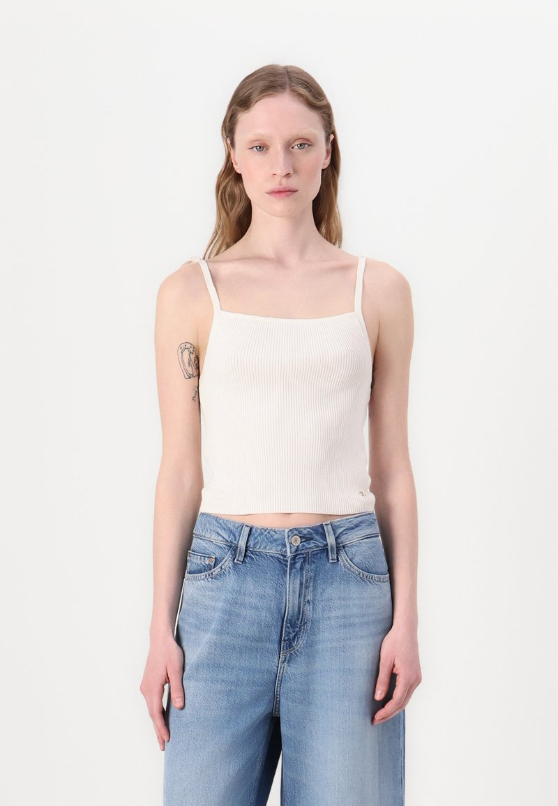 Hedvábné bílé crop top s tenkými ramínky, spárované s světle modrými kalhotami širokého střihu. Hladká textura, obdélníkový výstřih.