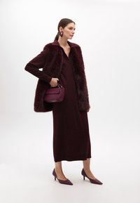Burgundská pletená šaty s výstřihem do V, vrstvená s umělou kožešinovou vestou. Hluboká burgundská kabelka a odpovídající střevíce na špičku dokončují vzhled.