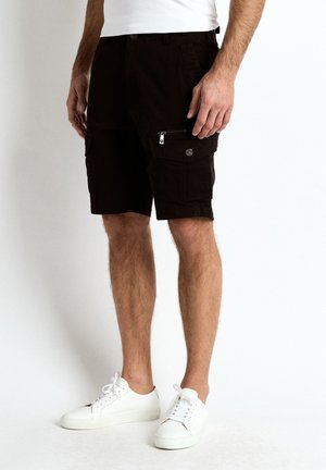 Mand iført sorte cargoshorts med lynlåslomme og hvide sneakers, stående mod en enkel lys baggrund.