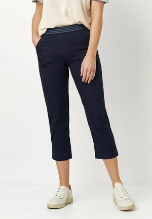 Navy Cropped-Hose mit einem strukturierten Bund, ausgestattet mit frontalen Taschen und geradem Schnitt. Kombiniert mit weißen Sneakers vor einem neutralen Hintergrund.