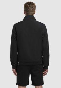Chaqueta negra con un alto cuello, puños elásticos y cinturilla. Presenta un diseño minimalista y una textura suave, acompañada de pantalones cortos negros.