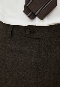 Pantaloni marroni con texture e chiusura a bottone, abbinati a una camicia bianca e una cravatta scura con strisce bianche.