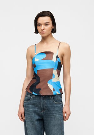 Rövid sötét hajú nő, aki egy testhezálló, kék, barna és bézs terepmintás pántos felsőt és kék farmernadrágot visel, egyszerű háttér előtt áll.