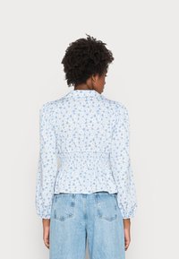 Blusa floral azul com mangas bufantes e cintura franzida, apresentando um tecido leve, colarinho e pequenos padrões circulares em branco.