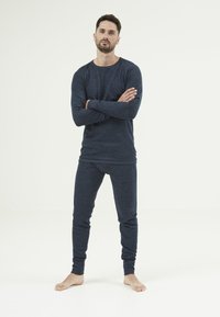 Tummansiniset pitkähihainen paita ja yhdistelemät housut pehmeästä, teksturoidusta kankaasta, kireillä hihoilla, jalat paljaina, руками ristissä.