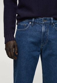 Jeans de mezclilla azul oscuro con un ajuste clásico, que cuentan con cinco bolsillos, costuras visibles y cremallera. Acompañados de un suéter oscuro.