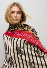 Bufanda de estampado de leopardo con rayas alternas y una banda roja con texto. Hecha de tela suave, se coloca de manera holgada alrededor del cuello.