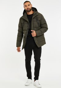 Olivenfarvet polstret jakke med hætte, lynlåslommer på brystet og sidelommer. Båret over en sort sweater og parret med sorte jeans og hvide sneakers.
