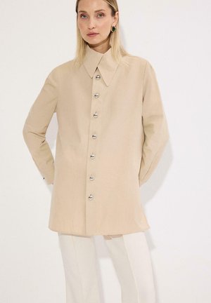 Femme aux cheveux blonds raides portant une chemise beige surdimensionnée avec des boutons argentés et un pantalon blanc, debout devant un fond blanc.