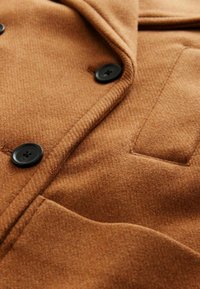Brun ullkappafabric med en mjuk textur, med svarta knappar och en bröstficka. Strukturerade slag framhäver designen.