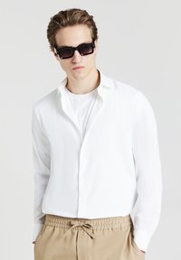 Camicia bianca a maniche lunghe con colletto aperto, abbinata a una maglietta bianca semplice e pantaloni beige con coulisse. Occhiali da sole neri con lenti scure.