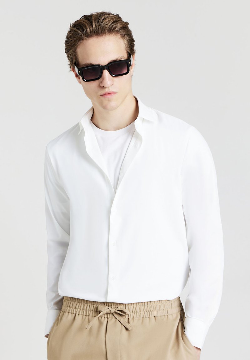 Camicia bianca a maniche lunghe con colletto aperto, abbinata a una maglietta bianca semplice e pantaloni beige con coulisse. Occhiali da sole neri con lenti scure.
