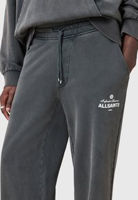 Grijze sweatpants van zachte stof, met een trekkoord in de taille, zijzakken en een witte geborduurde logo op de dij.