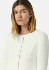 Weißes strukturiertes Jacket mit rundem Ausschnitt und ohne sichtbare Verschlüsse. Der Stoff wirkt gewebt mit einem dezenten Muster.