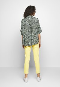 Žena s kudrnatými vlasy, oblečená v volné košili se vzorem, žlutých kalhotách a bílých teniskách, stojí čelem ke jednoduché bílé zdi.