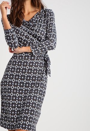 Robe à motif noir et blanc avec des manches longues, un décolleté drapé et une taille nouée, fabriquée en tissu doux avec une texture lisse.