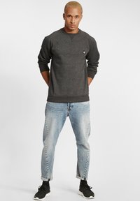 Donkergrijze sweatshirt met ribgebreide manchetten en zoom, voorzien van een klein logo op de borst. Draagt in combinatie met lichtblauwe jeans en zwarte sneakers.