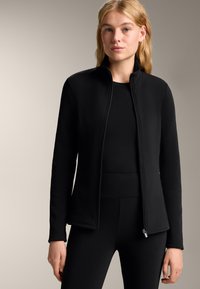 Czarna kurtka na zamek z wysokim kołnierzem, dopasowany krój, miękka tekstura i boczne kieszenie, zestawiona z dopasowanym czarnym topem i legginsami.